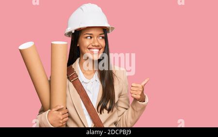 Beautiful hispanic woman holding paper blueprints pointing thumb up to the side smiling happy with open mouth Stock Photo