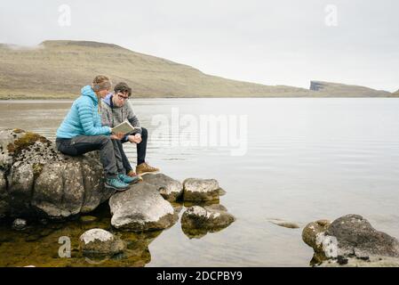 Young man and woman sitting on stones and reading book on gray day near sea Stock Photo