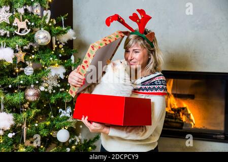 Happy mom with reindeers horns on the top of the head opening the gift with ragdoll cat by the Christmas tree and shining fireplace Stock Photo