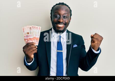 Handsome young black man wearing business suit holding 10 colombian pesos screaming proud, celebrating victory and success very excited with raised ar Stock Photo