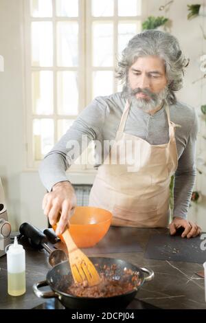 Serious mature man with beard mixing minced meat in frying pan by electric stove Stock Photo