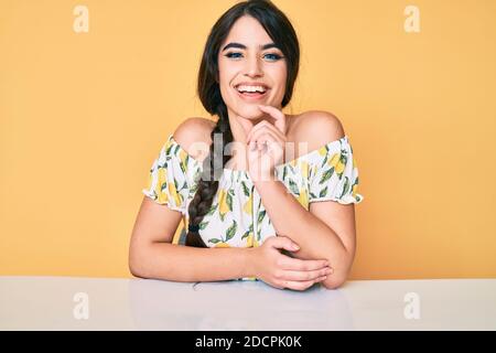 Brunette teenager girl wearing casual clothes sitting on the table looking confident at the camera with smile with crossed arms and hand raised on chi Stock Photo