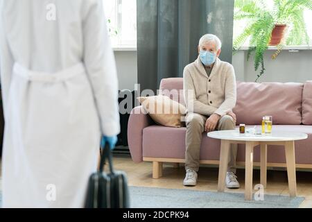 Sick senior man in casualwear and protective medical mask sitting on couch Stock Photo