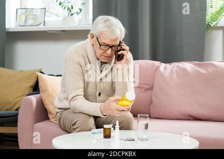 Senior man in casualwear sitting on couch, holding pills and calling doctor Stock Photo