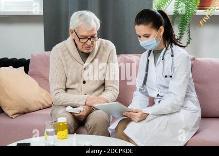 Young female doctor in whitecoat and protective mask using digital tablet Stock Photo