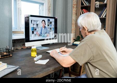 Senior man writing down advice of doctor while sitting in front of computer Stock Photo