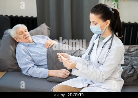 Senior man lying on couch next to young doctor with electronic thermometer Stock Photo