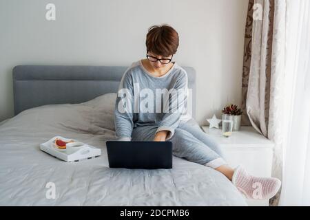 Caucasian woman in glasses and casual home clothes using laptop sitting on her bed and having breakfast. Remote working from home. Time at home during Stock Photo