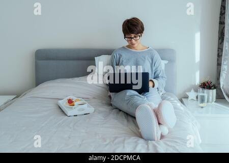 Caucasian woman in glasses and casual home clothes using laptop sitting on her bed and having breakfast. Remote working from home. Time at home during Stock Photo