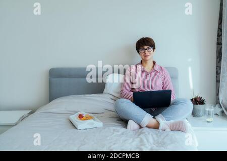 Caucasian woman in glasses, shirt and pajamas trousers using laptop sitting on her bed and having breakfast. Appropriate look for remote working from Stock Photo
