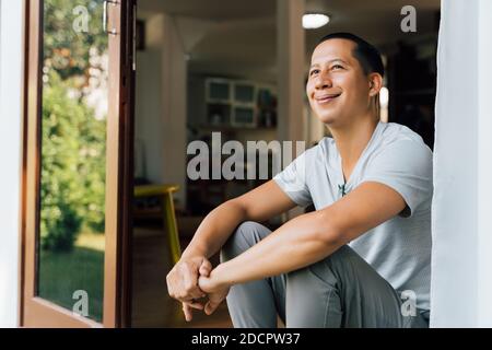 Portrait of 30-40 year happy smiling and thoughtful man sitting on the front door at home in summer. Mixed race of Asian and Caucasian guy looking away - with copy space Stock Photo