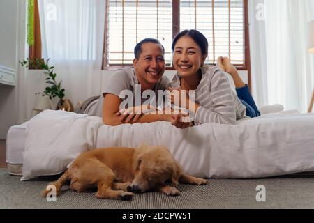 Portrait of happy young adult Asian couple looking at camera on bed together with dog pet in bedroom interior scene. 30s mature husband and wife smiling with happiness. Happy relationship life concept Stock Photo