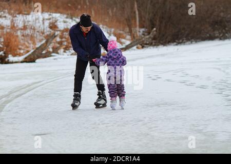 A father teaches little girl to skate on a frozen lake on a cloudy winter day Stock Photo