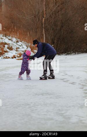 A father teaches little girl to skate on a frozen lake on a cloudy winter day Stock Photo