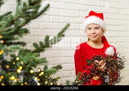 Girl in a red gnome hat hangs a garland on a christmas tree. installing a christmas lights on a christmas tree. decorations for christmas Stock Photo