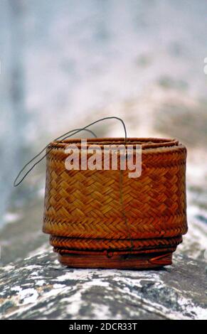 A handmade bamboo sticky rice container sits on a wall ledge in Luang Prabang, Laos.  The container is used to carry and keep the rice warm and moist. Stock Photo