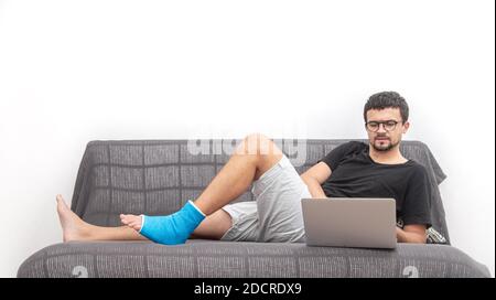 Man with glasses with broken leg in blue splint for treatment of injuries from ankle sprain working on a laptop on couch at home. Stock Photo