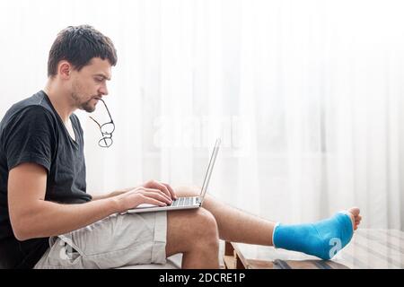 Young man with a broken leg in blue splint for treatment of injurie and ankle sprain is using a laptop at home. Stock Photo