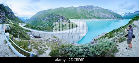 Beautiful panoramic scenery of clear lake with blue water, green hills and mountains around. Man taking photo using smartphone. Asphalt road along Lac de Moiry. Stock Photo