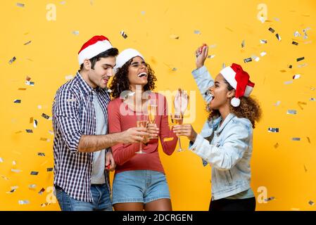 Group of three happy diverse friends celebrating Christmas drinking champagne on yellow studio background with confetti Stock Photo