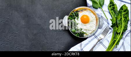 Fried eggs with spinach on iron pan plate. Fried eggs breakfast with white towel and spinach leaves, ingredients on dark black concrete background Stock Photo