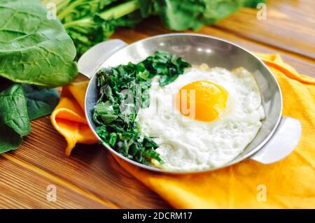 Green cooked spinach with fried egg on iron pan plate. Fried eggs healthy breakfast on wooden table background on orange napkin with spinach leaves Stock Photo