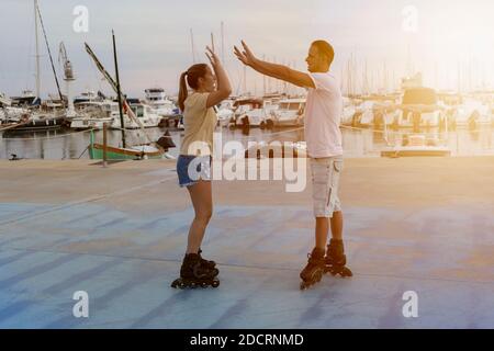 Young beautiful couple roller skating giving high five two hands at sunset in skate park next to the port in summer.Friendship lifestyle concept, succ Stock Photo