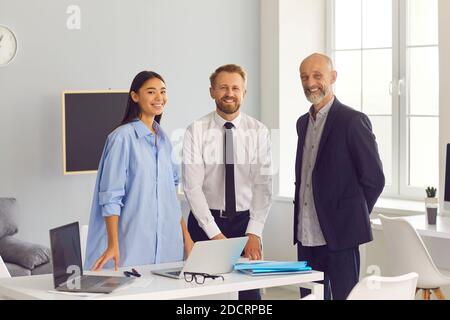Diverse team of smiling company employees standing near desk with laptops looking at camera Stock Photo