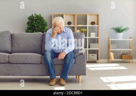 Senior man lowers his head and touches his hand to his forehead while sitting on the couch at home. Stock Photo