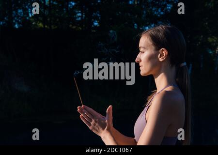 Yoga woman in sport clothes meditate and breathe on a sunrise in nature. Smoke from aromatic stick in hands. Stock Photo