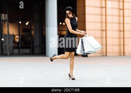 Cheerful stylish happy girl, in a black dress and hat, loves to go shopping: she carries shopping bags and runs to the Mall for the latest offers. Sho Stock Photo