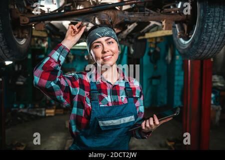 Portrait of a young happy female mechanic in uniform posing with tablet at her hands and remove glasses from the head. The car is located on the lift. Stock Photo