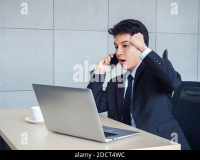 Young handsome Asian businessman in suit raising fist with exciting and shocking while calling with his mobile phone and looking laptop computer on de Stock Photo