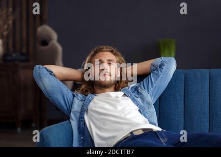 Time to relax. A curly-haired guy in a casual style, sitting on the couch at home, smiling and relaxing. Pleasant leisure time. Rest at home Stock Photo