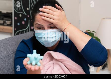 Concerned adult woman wearing surgical or medical mask having sars covid flu symptoms holding plastic virus replica in hand as pandemic concept Stock Photo