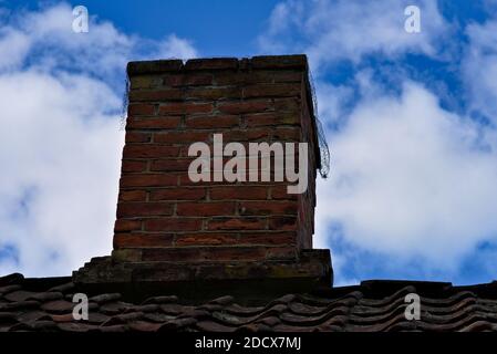 Old classic clay brick chimney on a rooftop with rooftiles that looks worn and weathered. Stock Photo