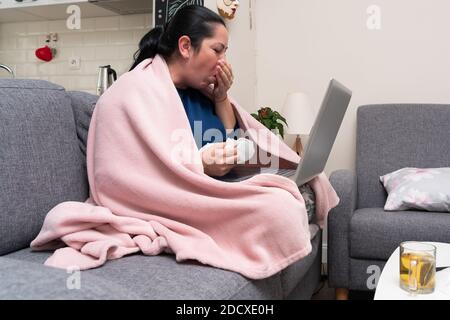 Ill adult female model coughing as working remotely from home with personal laptop on couch with tea on table feeling unwell having influenza sars cov Stock Photo