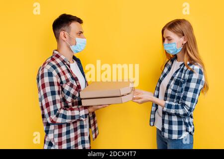 man and a woman in checked shirts and medical protective masks on their faces, holding pizza boxes on an isolated yellow background. Stock Photo