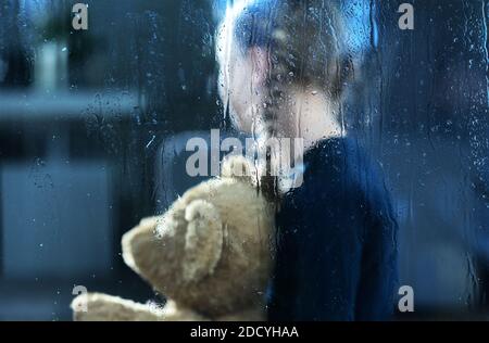 Girl with Teddy Bear Behind Window Covered by Rain Drops. Children Depression and Social Issues Conceptual Photo. Stock Photo