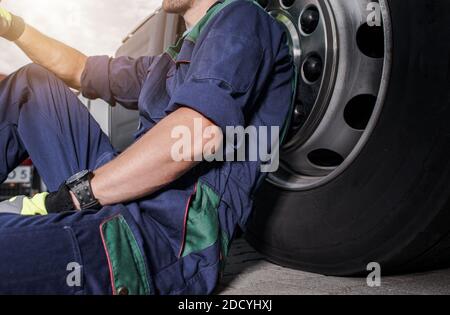 Caucasian Heavy Duty Truck Mechanic in His 40s Replacing Semi Truck Tires Theme. Seating Next to Vehicle Wheel. Stock Photo