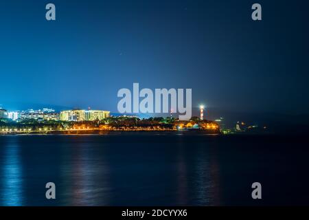 Sea night landscape with cape, illuminated buildings and lighthouse. Stock Photo