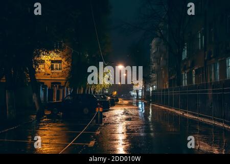 Empty city street in rainy weather at night illuminated by city lamps, no people, wet and puddles with reflection, horror and mystery atmosphere. Stock Photo