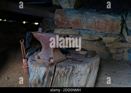 Old abandoned blacksmiths workshop with rusted anvil, hammer and tools on a worn wood log. Stock Photo