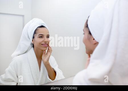 Portrait of beautiful mixed race woman reflected in the mirror wearing bathrobe touching clean, fresh skin of her face after washing Stock Photo