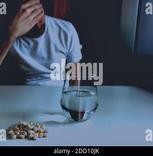Depressed man considering suicide by pill overdose, sitting at white table with intentional blurred blacked out background Stock Photo