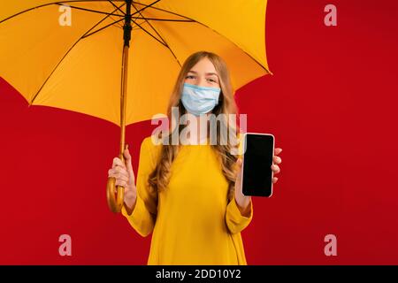 A beautiful young womanin a protective mask on her face, with an umbrella in her hands, shows her mobile phone Stock Photo