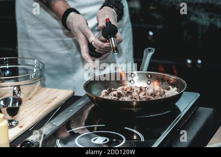 The chef cooks small octopus on a metal frying pan at the restaurant to make brazilian dish. Pan is on fire. Stock Photo