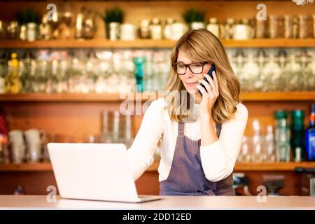 Shot of smiling female waitress using mobile phone and laptop while standing at the counter and working in the cafe. Small business. Stock Photo