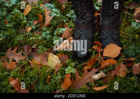 Hello autumn. Closeup on woman standing in boots on the green grass with autumn leaves outdoors in the city park in autumn. Stock Photo