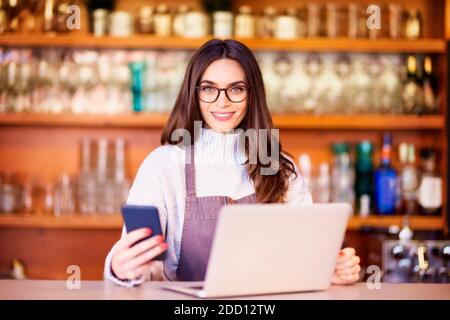 Portrait of a beautiful waitress text messaging and using laptop while standing behind the counter at a restaurant. Small business. Stock Photo